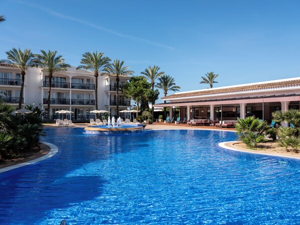 A large swimming pool with lounge chairs and umbrellas, surrounded by palm trees and a multi-story building with balconies.