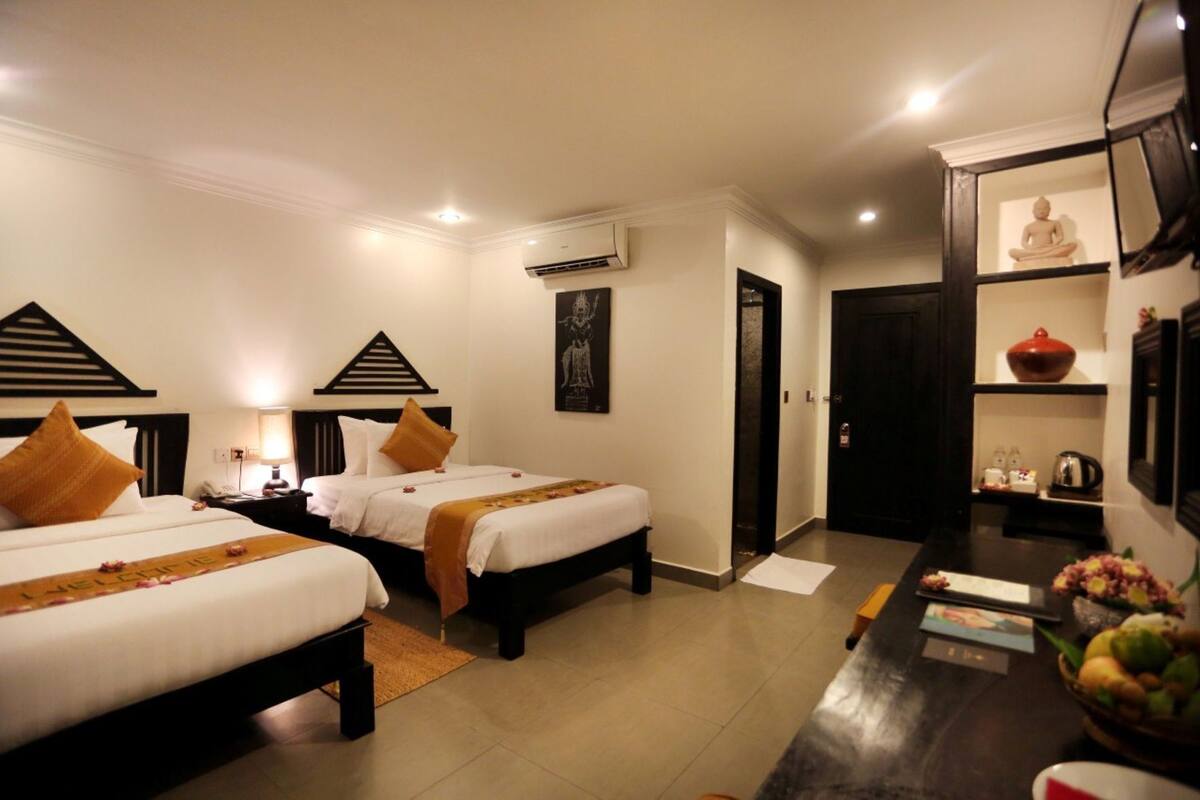 A hotel room with two beds, a desk, a chair, a small table, a wall-mounted air conditioner, a framed picture, and a shelf with decorative items.