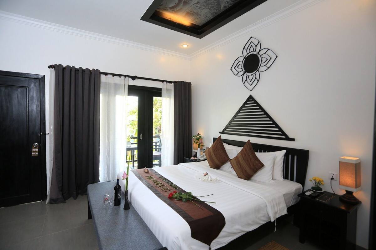 A hotel room with a bed, a bedside table with a lamp, a wall-mounted decorative piece, and a view of the outdoors through a door.
