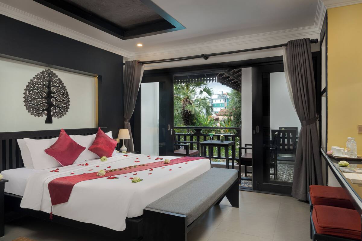 A hotel room with a bed, a bench, a desk, and a balcony with a view of greenery.
