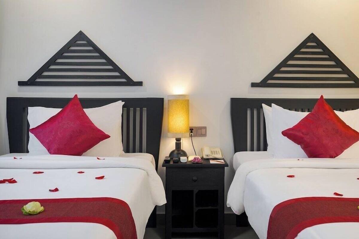 A hotel room with two beds, each with a red pillow, a bedside table with a lamp, and a small nightstand with a phone.