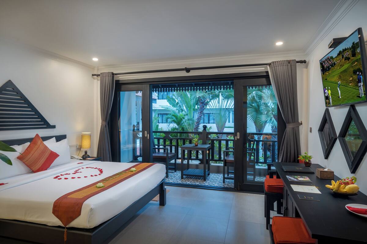 A hotel room with a bed, a desk, and a balcony with a view of palm trees and a building.