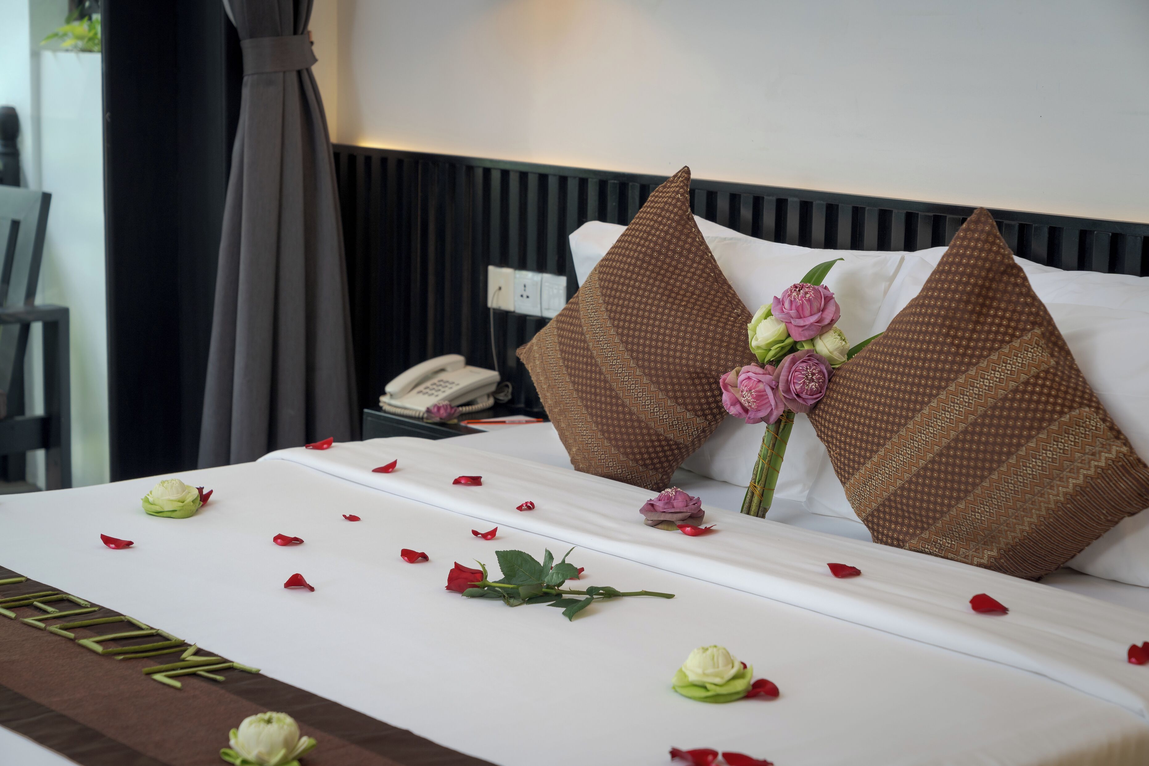 A neatly made bed with rose petals and a bouquet of flowers, a telephone, and a dark curtain in a hotel room.