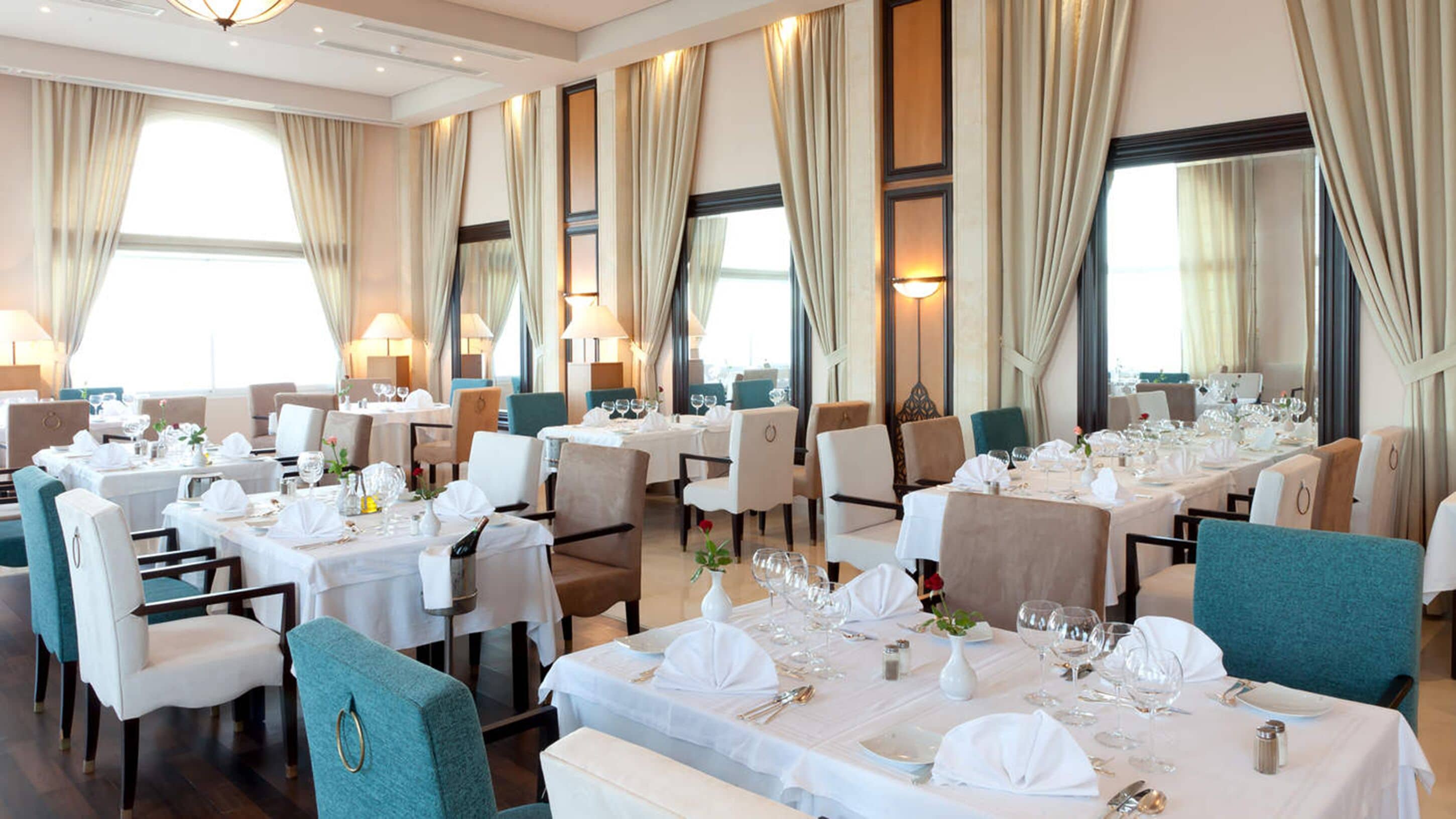 A spacious dining area with multiple tables set for dining, large windows with curtains, and a modern chandelier.