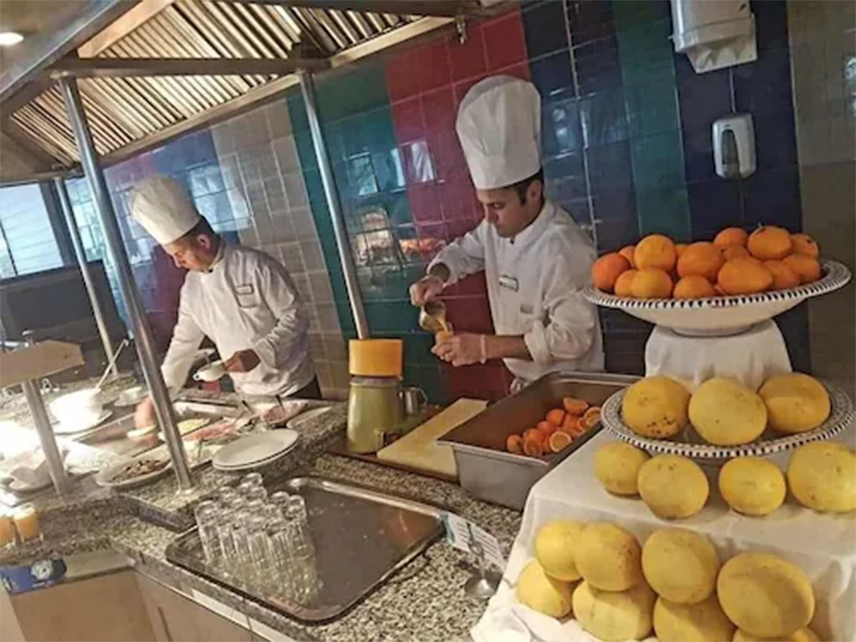 Een keuken met koks die eten bereiden, een buffet met fruit, en een spoelbak met glazen.