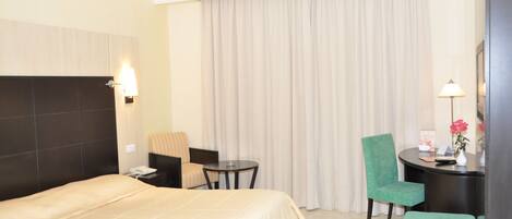 Quarto de hotel com uma cama grande, uma escrivaninha com cadeira, uma mesinha e um banquinho verde.
