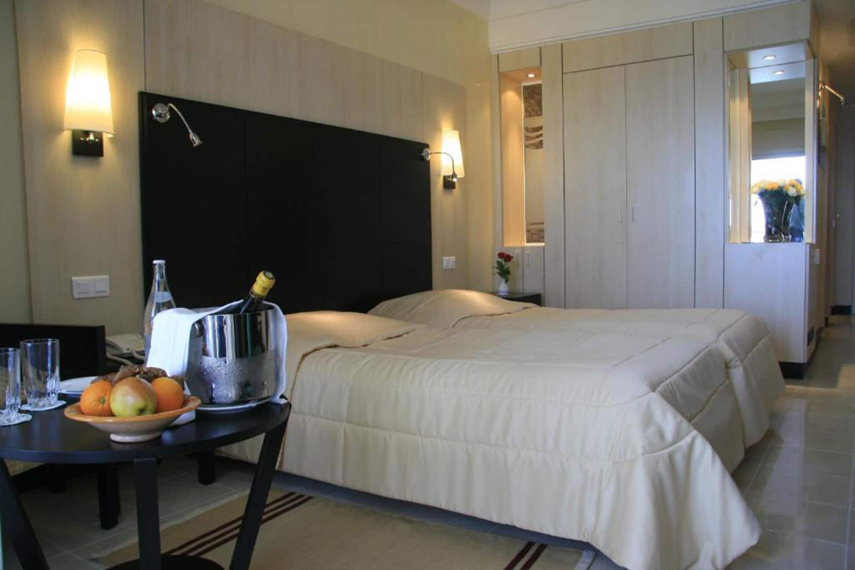 Une chambre d’hôtel avec un lit, une table de chevet sur laquelle se trouve un panier de fruits, une bouilloire et une bouteille d’eau. Il y a un miroir au mur et une porte qui donne sur une autre pièce.