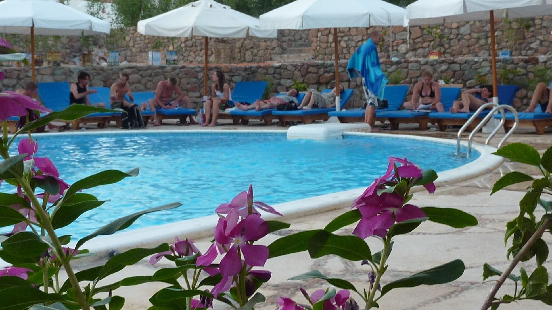Una piscina con sillas de descanso y sombrillas, rodeada de un muro de piedra y vegetación.