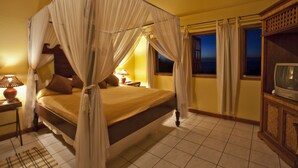 A four-poster canopy bed with a wooden headboard, a bedside table with a lamp, and a television on a wooden cabinet. - Villa Caprí (Gros Islet)
