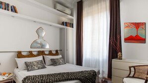 Habitación de hotel ordenada con cama, mesita de noche y una silla. Se observa una estantería con libros y un cuadro en la pared.