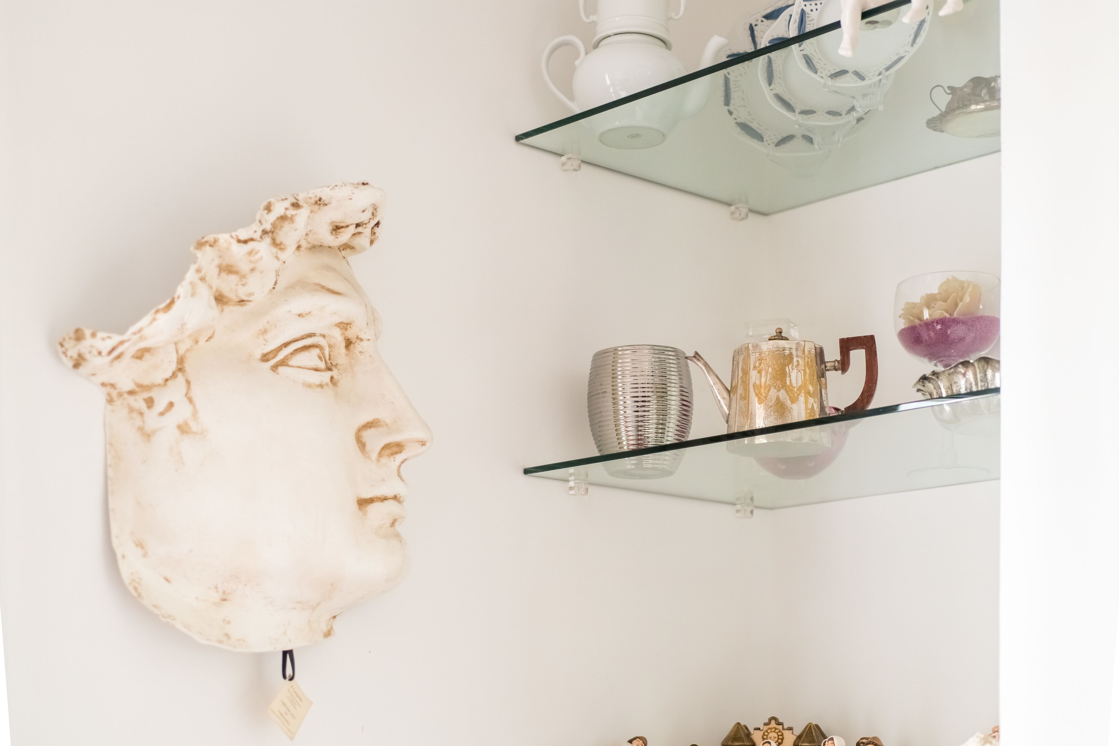A wall-mounted sculpture of a human face, a glass shelf with decorative items, and a white wall.