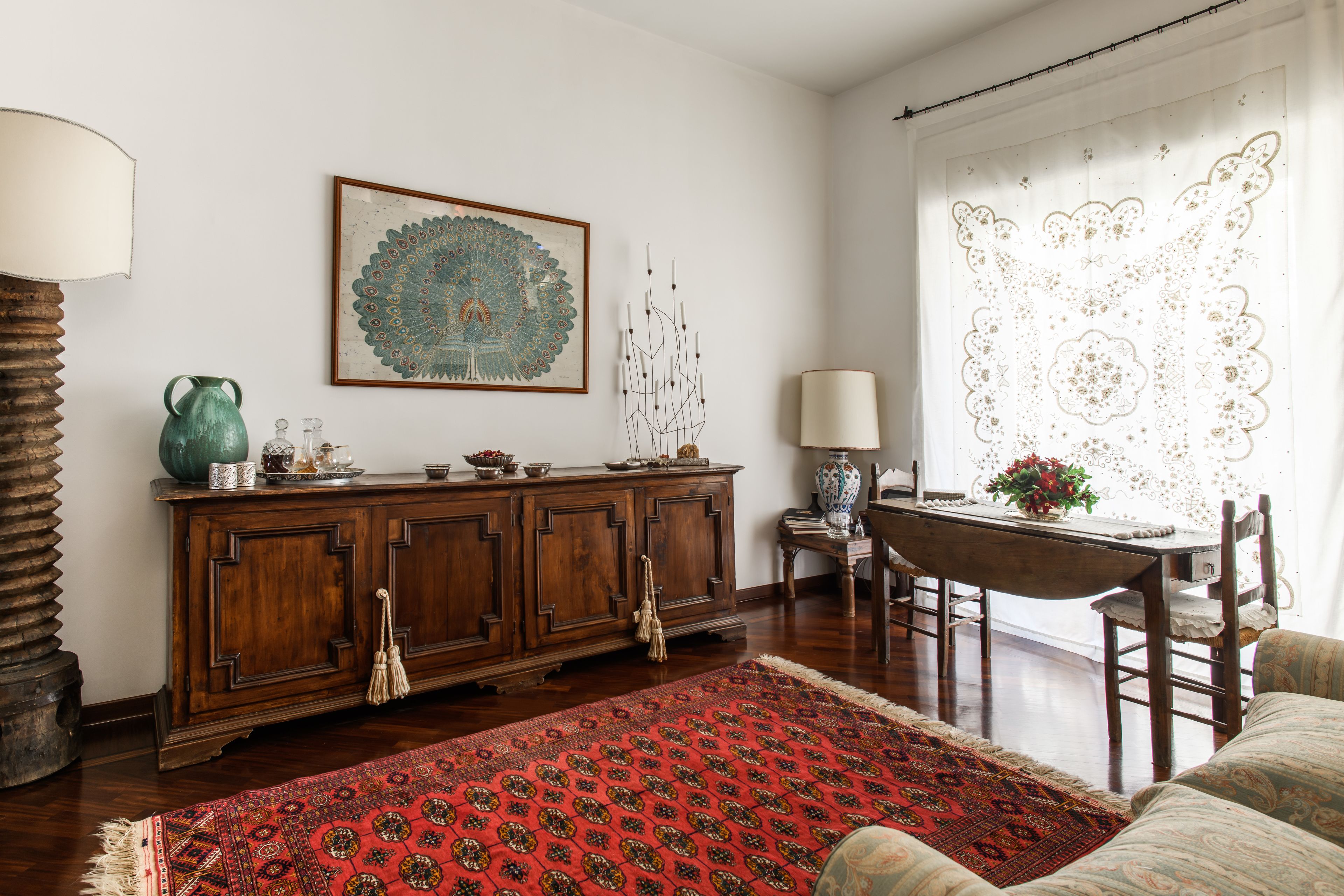 Un salon traditionnel avec un meuble en bois, un tapis rouge à motifs, une table à manger avec des chaises et un tableau décoratif représentant un paon accroché au mur.