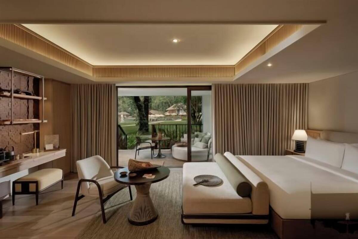 Une chambre d’hôtel moderne, dotée d’un canapé, d’une table ronde, d’un petit bureau et offrant une vue sur un jardin grâce à de grandes fenêtres.