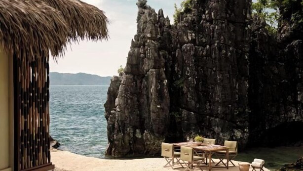 A beachside dining area with a thatched-roof structure, a table set for four, and a rocky cliff backdrop. - Lagen, El Nido (El Nido)