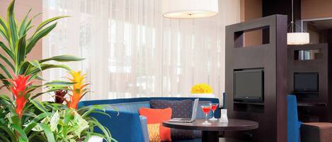 Una sala de estar moderna con un sofá azul, una mesa redonda con una computadora portátil y bebidas, un televisor montado en la pared y un ventanal con cortinas translúcidas.
