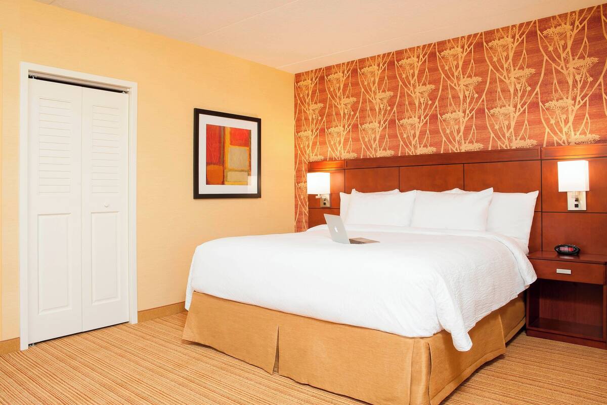 Habitación de hotel con cama, escritorio, lámpara, un cuadro enmarcado y una puerta.