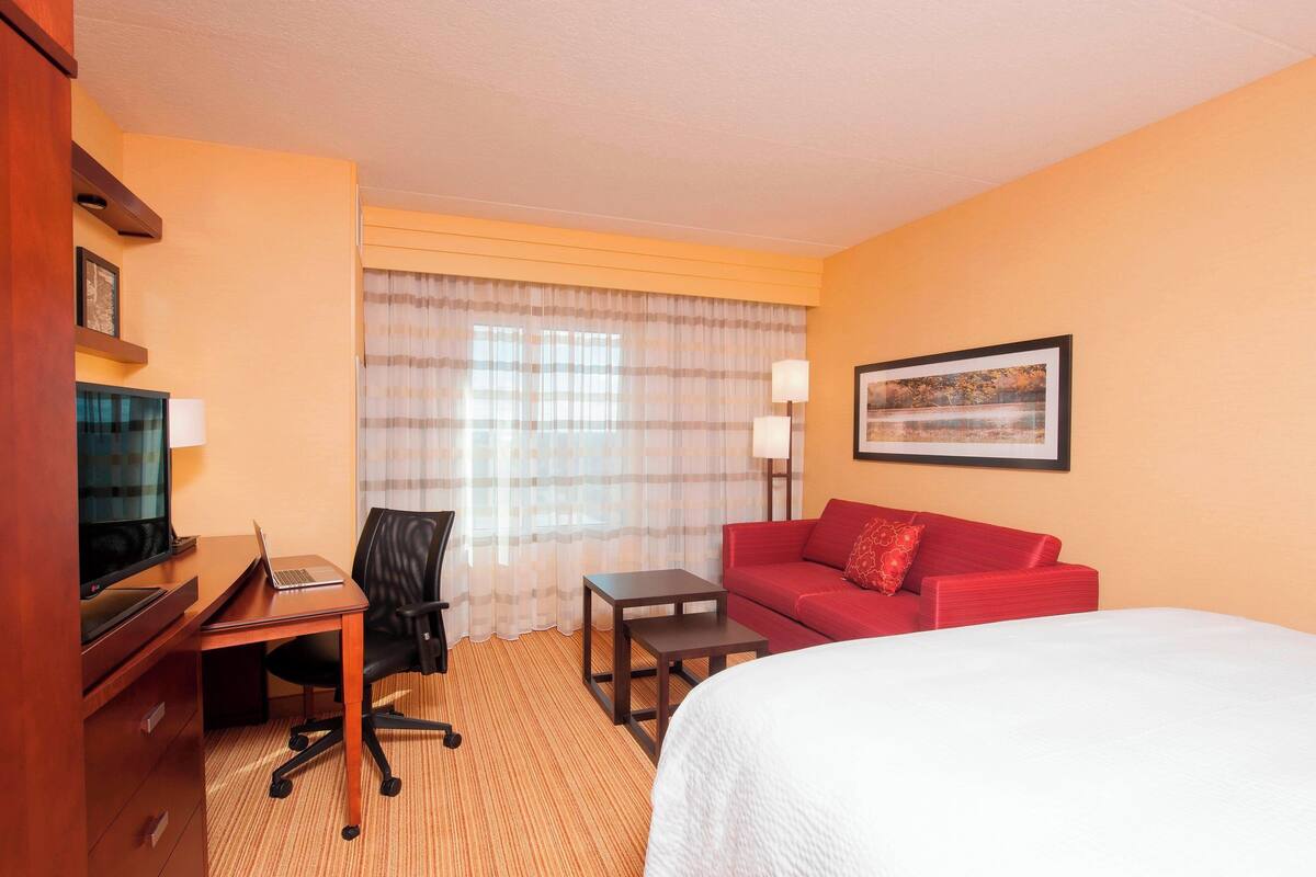 Habitación de hotel con cama, escritorio con computadora portátil, silla, sofá rojo, mesita pequeña y televisor.