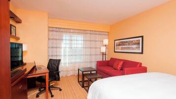 A hotel room with a bed, a desk with a laptop, a chair, a red sofa, a small table, and a TV.