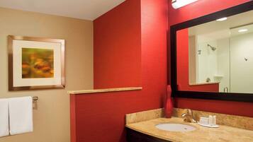 Un baño con pared roja, un cuadro enmarcado, lavamanos con artículos de aseo y un espejo que refleja una ducha.
