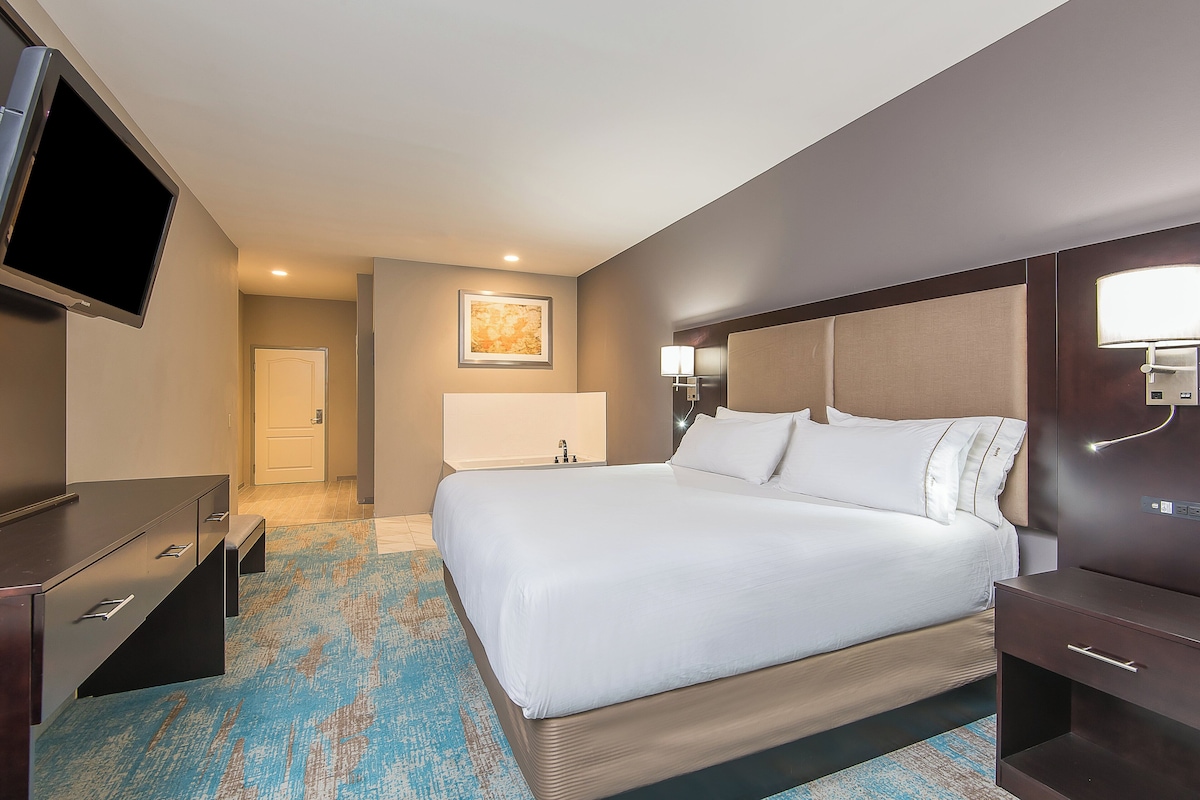 Habitación de hotel con cama, escritorio, televisor, lámpara de noche y puerta.