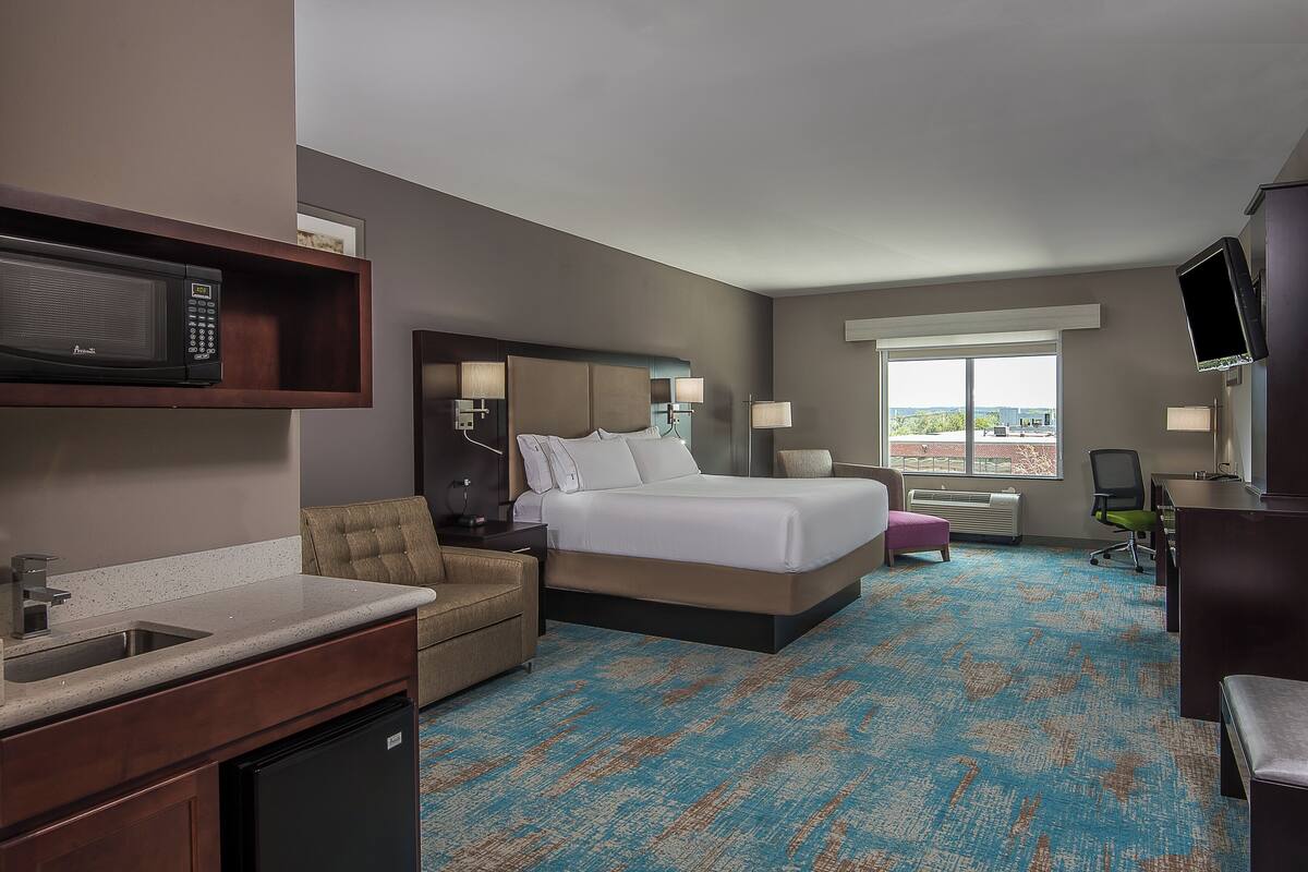 A hotel room with a bed, a desk, a chair, a TV, a microwave, a sink, and a window.