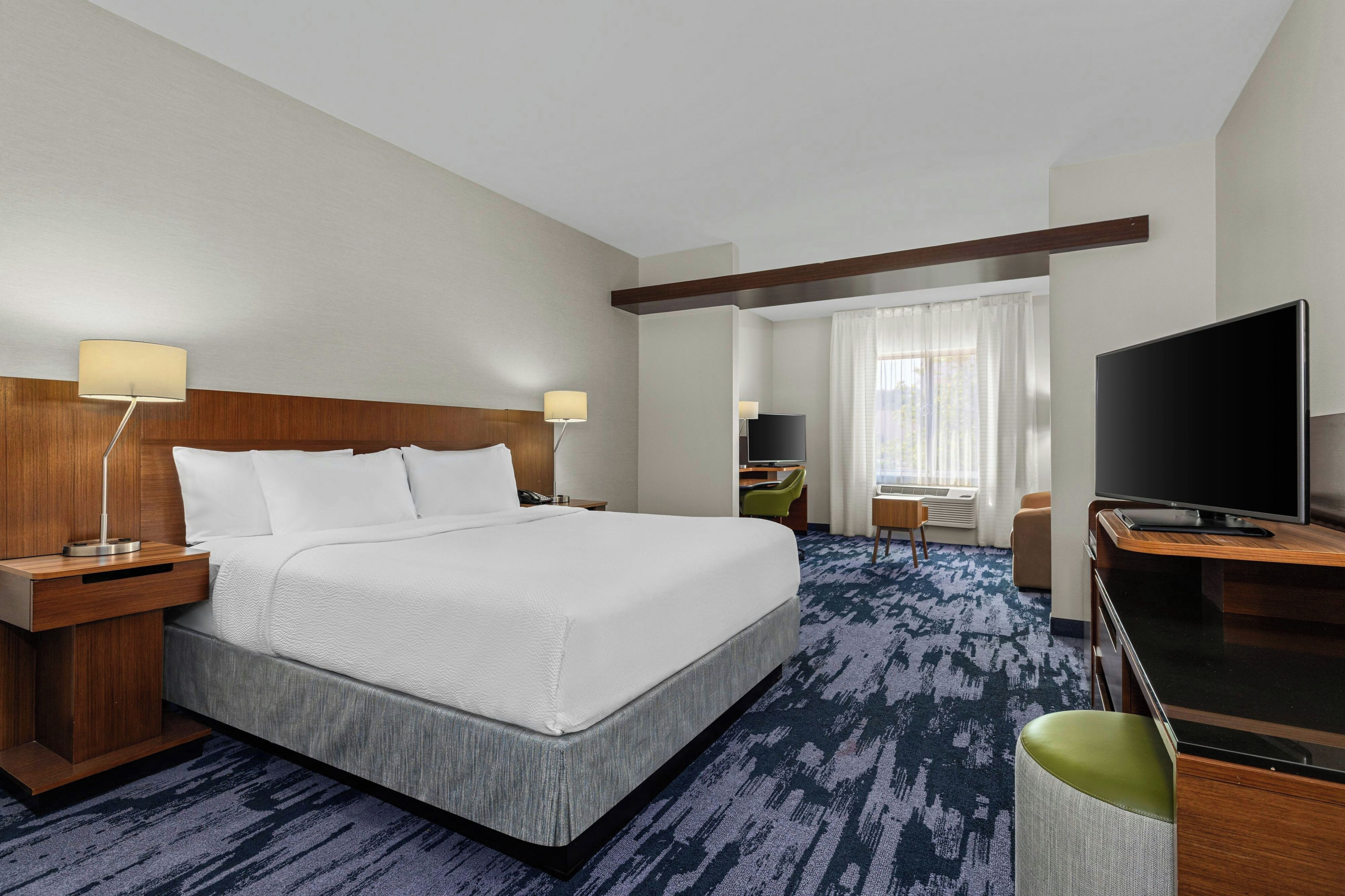 A hotel room with a large bed, a desk with a flat-screen TV, a chair, and a window with curtains.