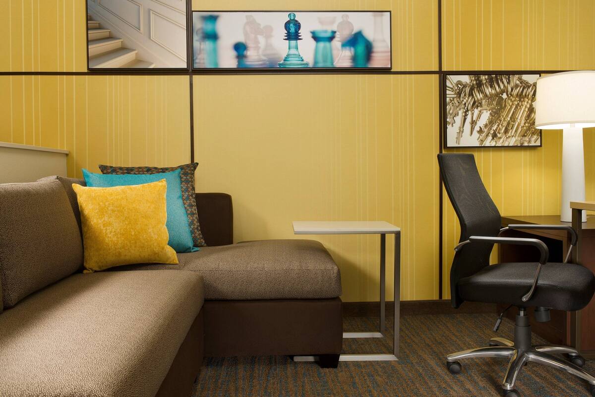 A hotel room with a sectional sofa, a small table, a chair, and a lamp.