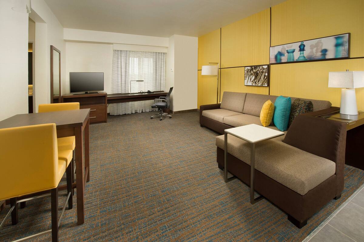 A hotel room with a sofa, a small table, a TV, and a desk with a lamp.