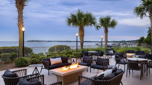 Un patio al aire libre con mobiliario de mimbre, una chimenea central y vistas al agua.