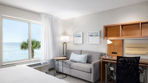 Habitación de hotel con una cama, un sofá gris, un escritorio con una silla, una lámpara y cuadros en la pared.