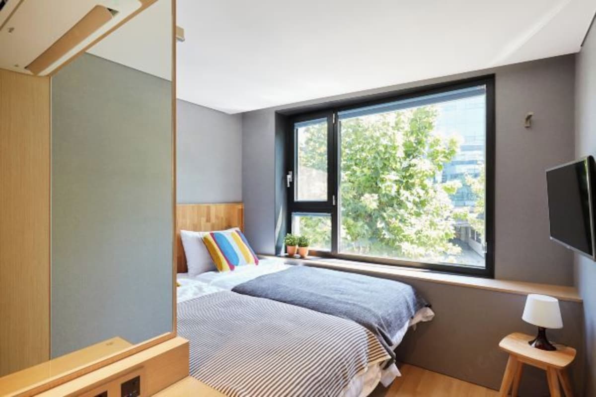 A modern bedroom with a large window, a bed with striped bedding, a bedside table, and a TV mounted on the wall.