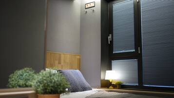 Une chambre d’hôtel avec un lit, une table de chevet, une lampe, une plante en pot et une fenêtre avec des stores.