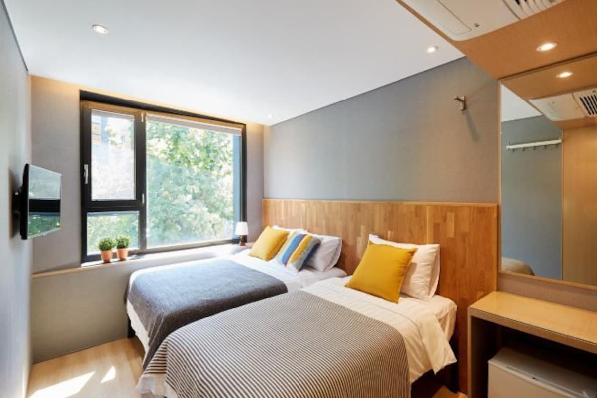 A modern hotel room with two beds, a large window, a mirror, and a small desk.