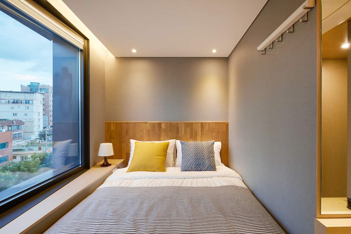 A modern hotel room with a large window, a wooden headboard, and a striped bedspread.