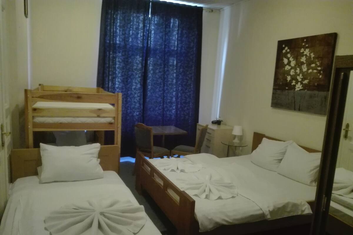 A hotel room with two single beds, a bunk bed, a small table, a chair, a desk, a lamp, and a painting on the wall.