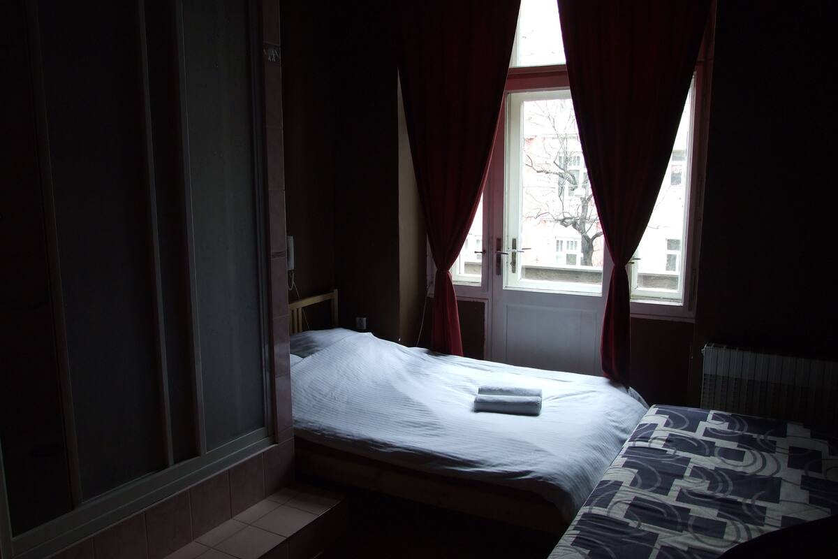 A small, dimly lit room with a single bed, a window with red curtains, and a radiator.