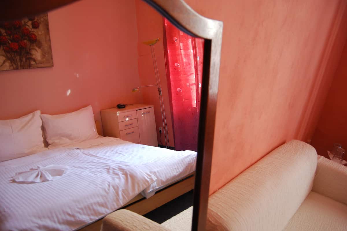 A neatly made bed with white linens and pillows, a bedside table with a lamp, and a pink wall with a floral painting.