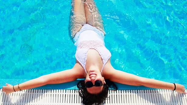 Una persona recostada en una tumbona junto a la piscina, con los brazos extendidos, flotando en una piscina azul cristalina.