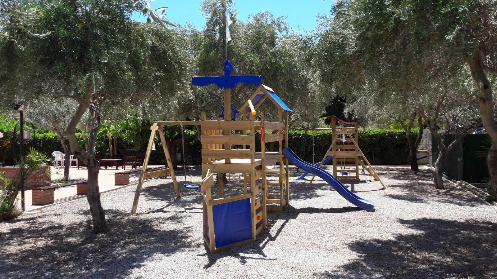 Un parque infantil con estructuras de madera para escalar y toboganes, rodeado de árboles y grava.