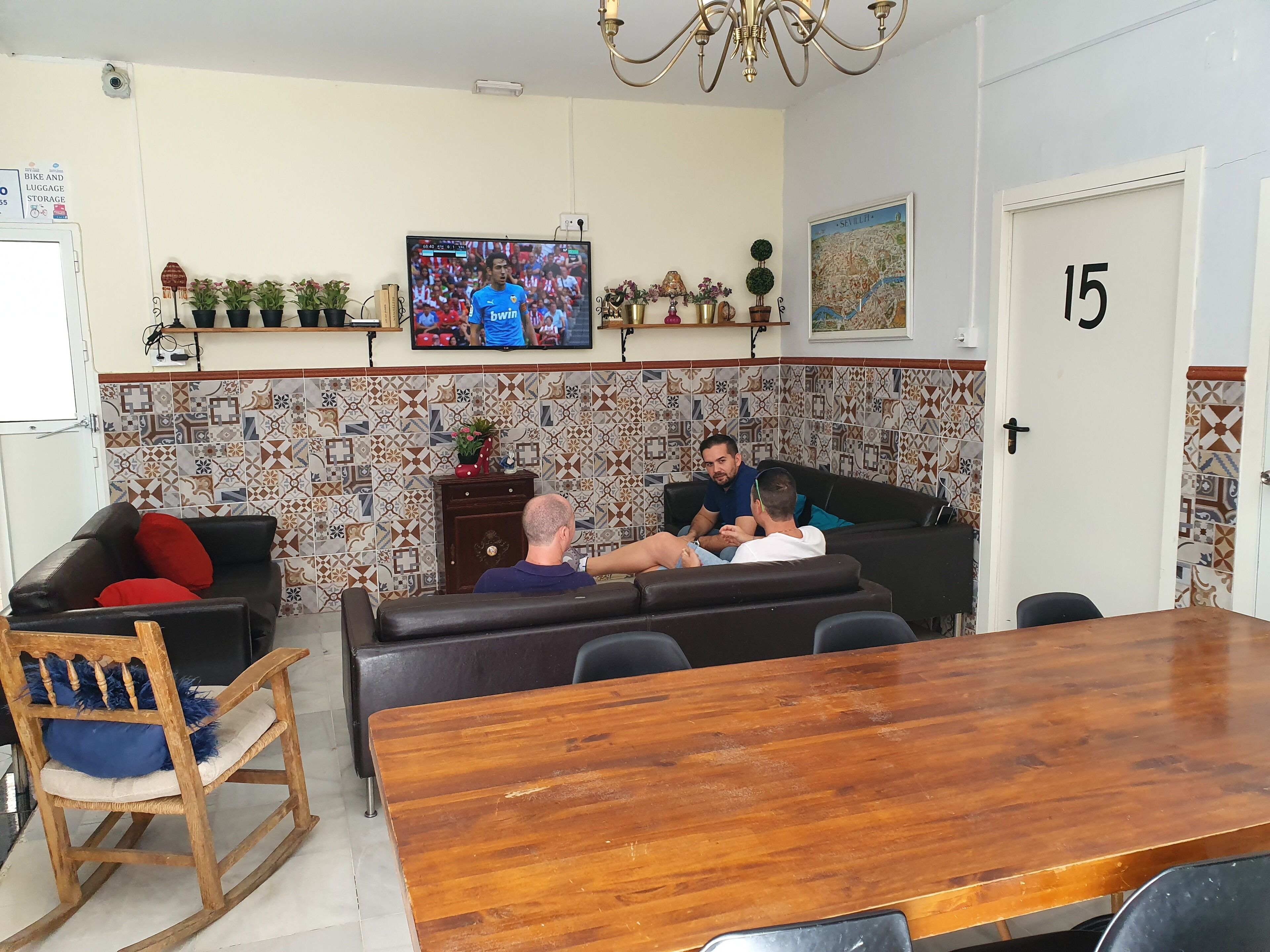 A living room with a television, a sofa, a wooden dining table, and a tiled wall.