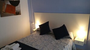 A neatly made bed with a patterned bedspread and two black pillows, a bedside table with a lamp, and a wall-mounted painting. - Sevilla Dream Hostel (Seville)