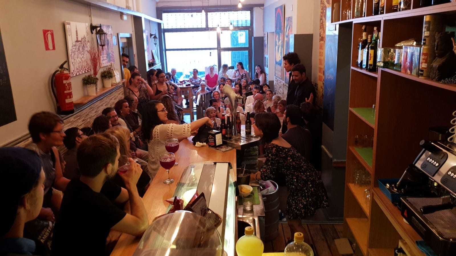 Zona de bar con clientes sentados en mesas, un camarero detrás de la barra y estanterías llenas de botellas.