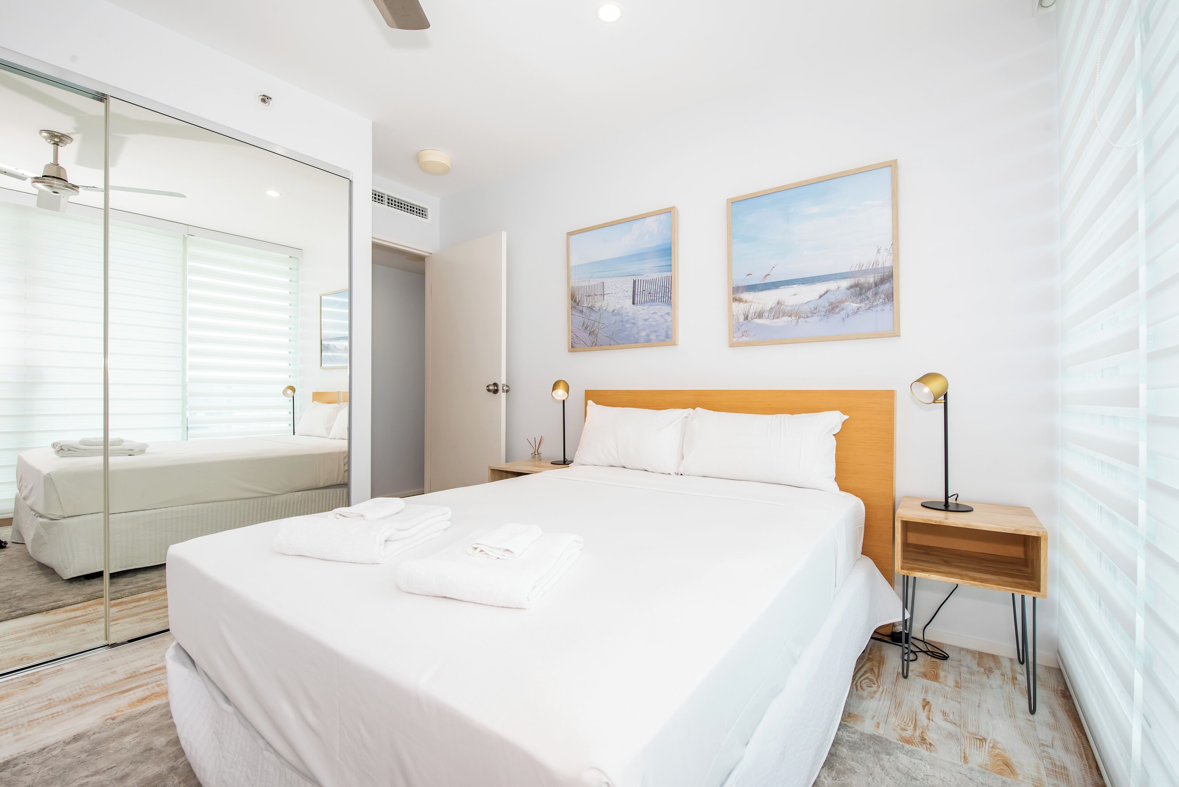 A modern hotel room with a large bed, a glass-enclosed walk-in closet, and two framed beach paintings on the wall.
