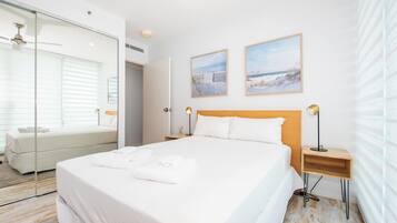 A modern hotel room with a large bed, a glass-enclosed walk-in closet, and two framed beach paintings on the wall.