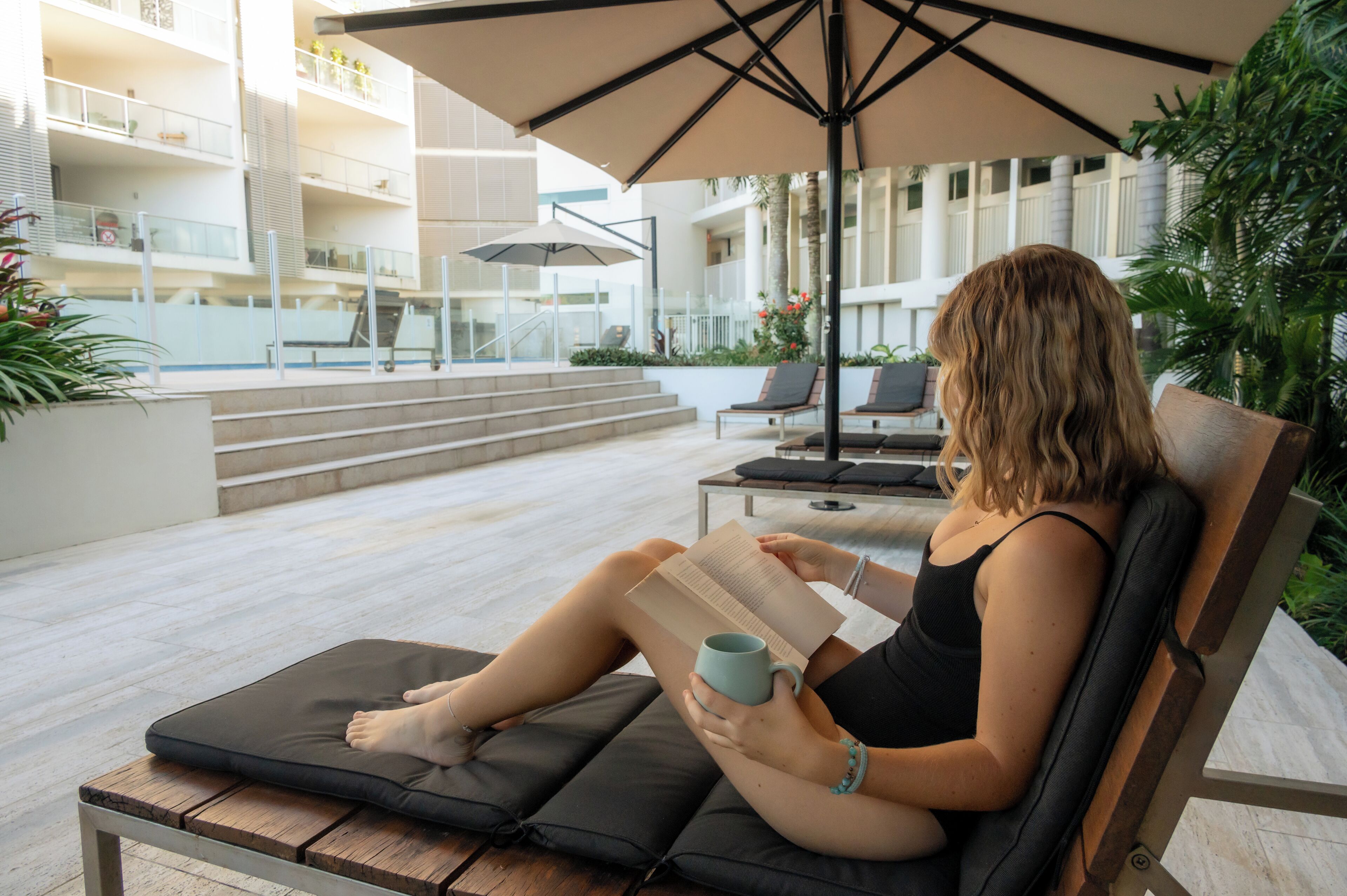 Eine Person sitzt entspannt in einer Liege unter einem Sonnenschirm, liest ein Buch und hält eine Tasse.