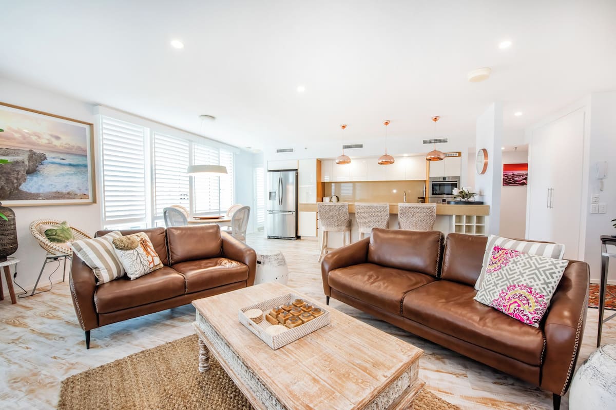 A modern living room with brown leather sofas, a wooden coffee table, and a kitchen in the background.