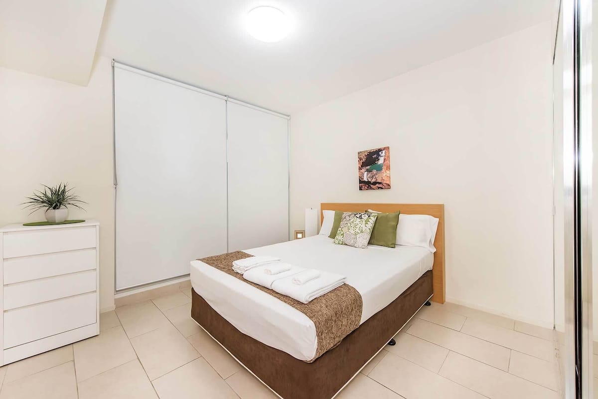 A bedroom with a bed, a bedside table, a potted plant, a painting, and a wardrobe.