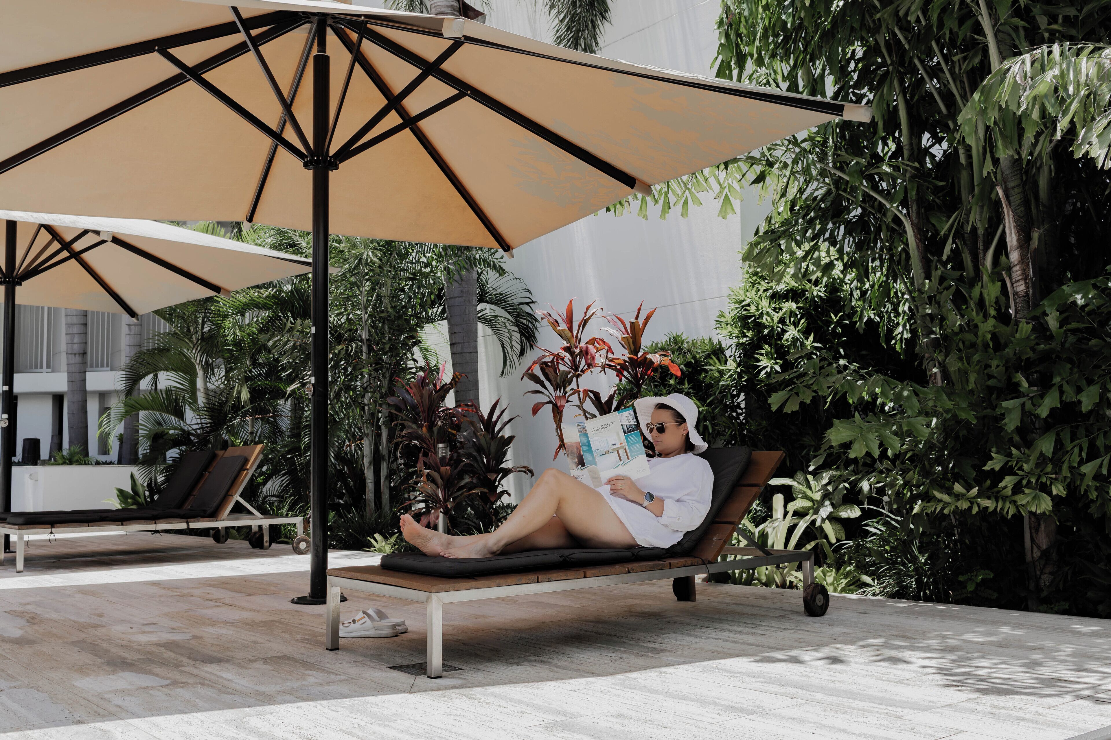 A person relaxing on a sun lounger under a large umbrella in a well-maintained outdoor area with various plants and a modern building in the background.