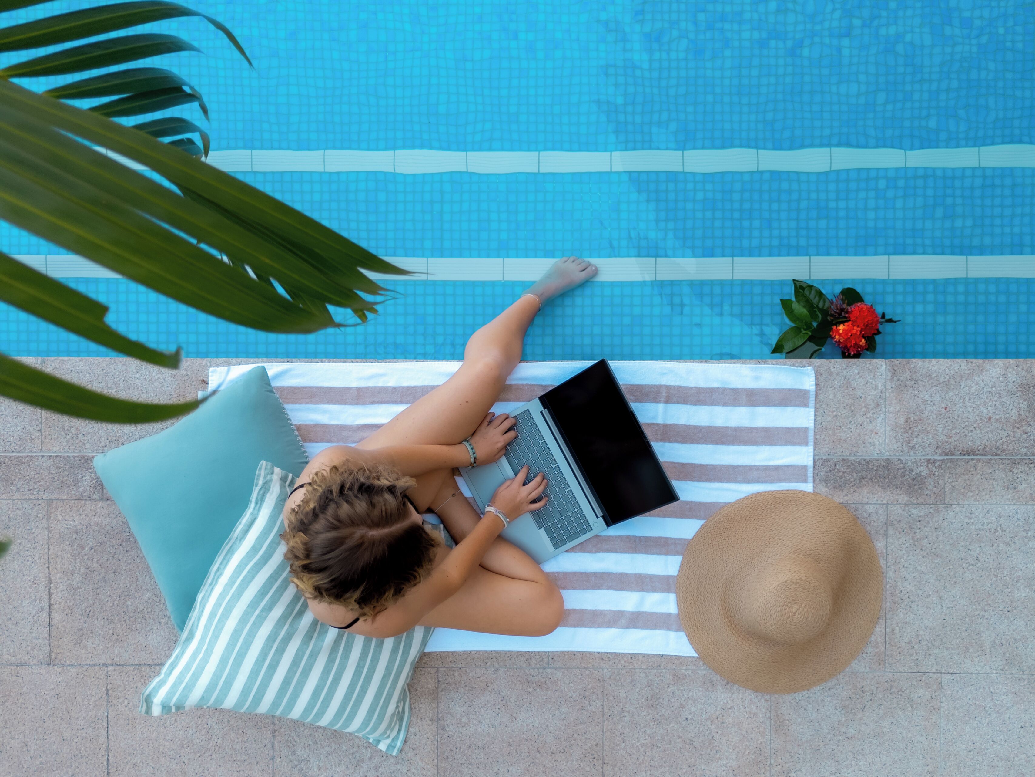Eine Person liegt auf einem gestreiften Handtuch am Pool und benutzt einen Laptop.