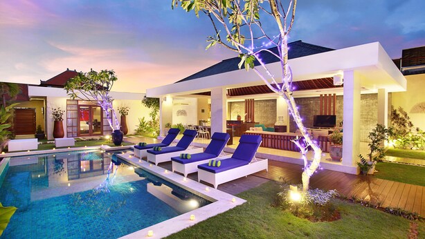 A modern outdoor pool area with lounge chairs, a bar, and a well-lit garden at dusk. - Indi Villas - The Ambika (Seminyak)